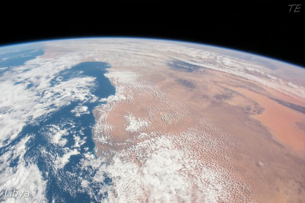 Blick auf Libyen aus dem Weltall mit Sahara-Wüste und Wolken über dem Mittelmeer, blauem Himmel und Erdkrümmung im Hintergrund.