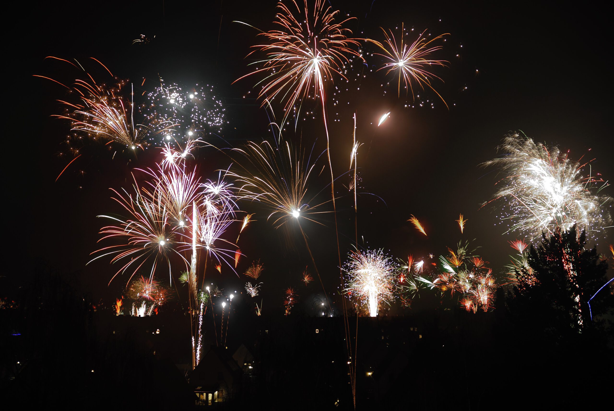 Leuchtende Feuerwerkskörper erhellen den Nachthimmel in verschiedenen Farben und Formen. Mehrere Feuerwerkskörper explodieren gleichzeitig und zeigen rote, gelbe, weiße und violette Lichtstrahlen. Einige der Feuerwerkskörper haben langgezogene Schweife, die sich in funkelnde Lichter auflösen, während andere in runden, strahlenden Mustern aufblühen. Am unteren Rand des Bildes sind dunkle Silhouetten von Häusern und Bäumen zu erkennen, die einen Kontrast zum hell erleuchteten Himmel bilden. Der gesamte Himmel ist mit Explosionen von Funken und Licht gefüllt, die eine festliche Atmosphäre vermitteln.