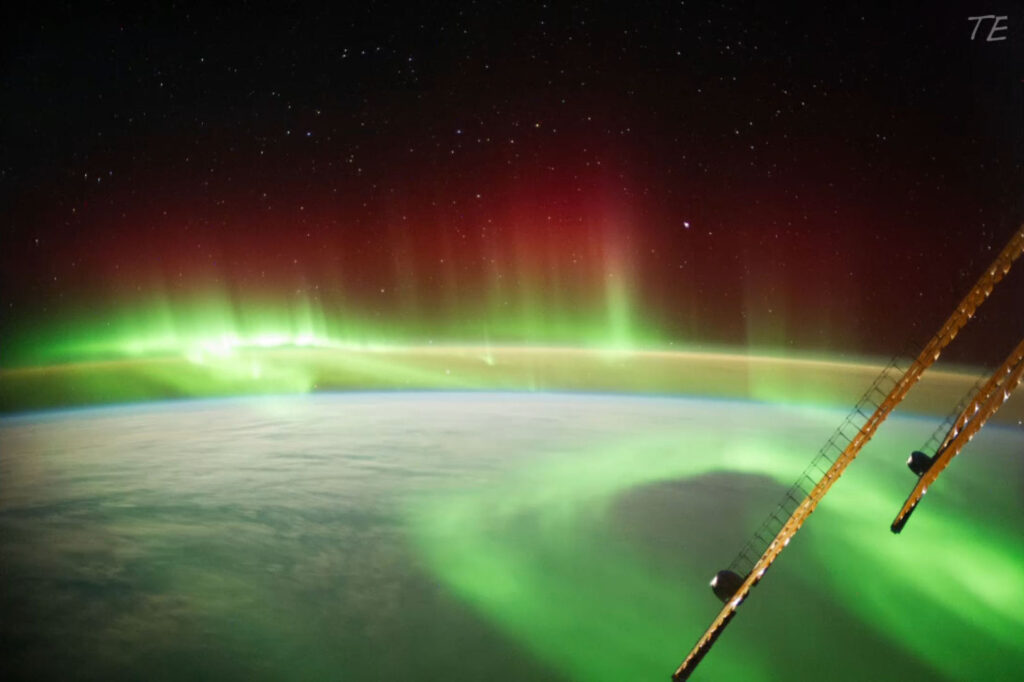 Blick aus der ISS auf die Erde mit leuchtenden Polarlichtern in Grün und Rot sowie Sternen am Nachthimmel.