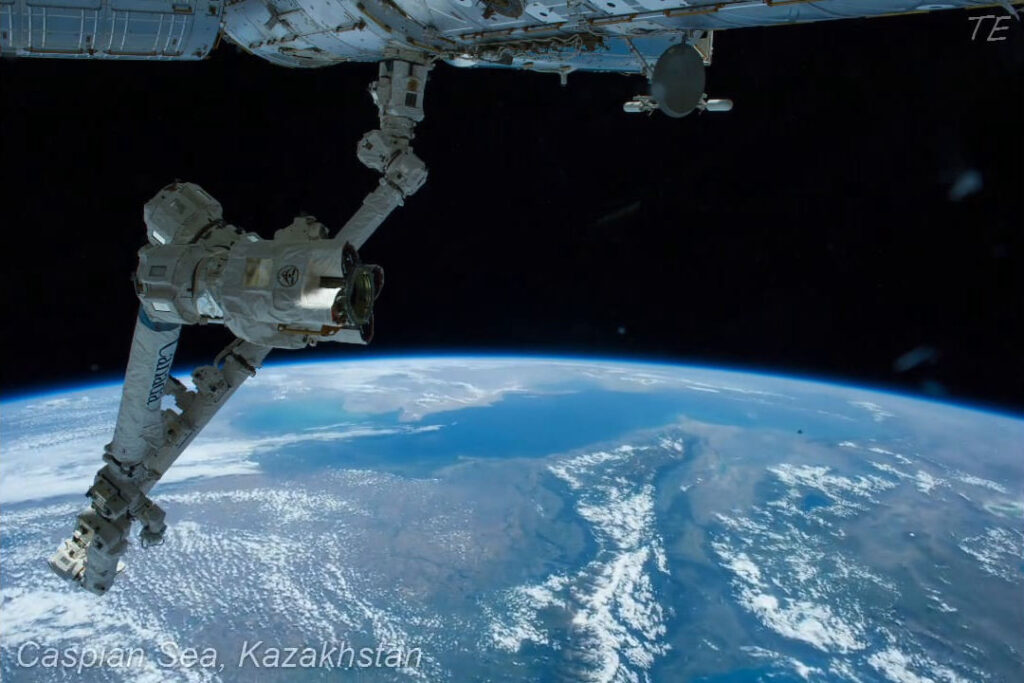 Blick aus der ISS auf die Erdoberfläche mit dem Kaspischen Meer und Kasachstan im Hintergrund, sowie einem Roboterarm im Vordergrund.