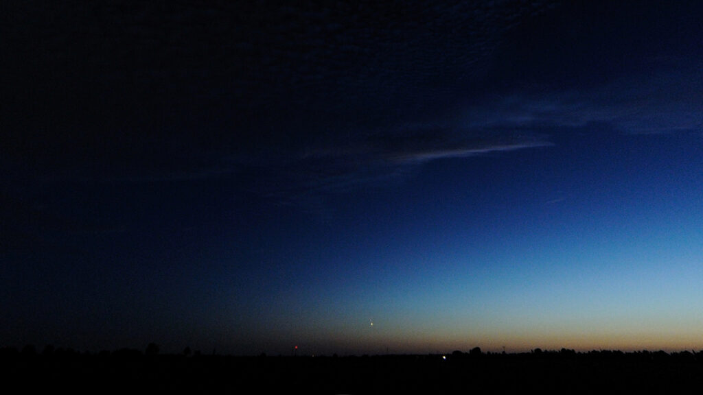 Dämmerungshimmel mit dunklen Wolken, Übergang von Blau zu Orange am Horizont, vereinzelte Lichter am Boden.