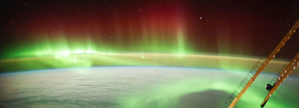 Blick aus der ISS auf die Erde mit leuchtenden Polarlichtern in Grün und Rot sowie Sternen am Nachthimmel.